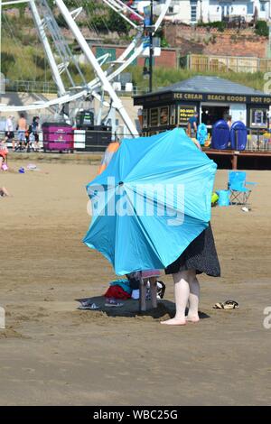 Donna che mette su un ombrello blu della spiaggia, Scarborough, North Yorkshire, Regno Unito, 26th agosto 2019, Tempo: Caldo e soleggiato agosto festa della banca Lunedi mattina e record di temperatura sono attesi per essere rotto in tutta l'Inghilterra orientale per la fine estate vacanza. La gente si affolla sul lungomare e sulla spiaggia da chilometri per divertirsi al sole in questa tradizionale cittadina balneare inglese sulla costa nord-orientale. Foto Stock