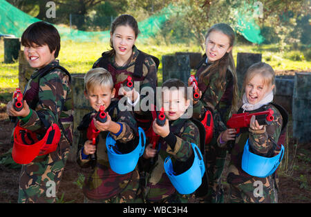 Gentile sorridenti gruppo di bambini paintball giocatori in camuffamento in posa con pistole di paintball sul campo da gioco all'aperto Foto Stock