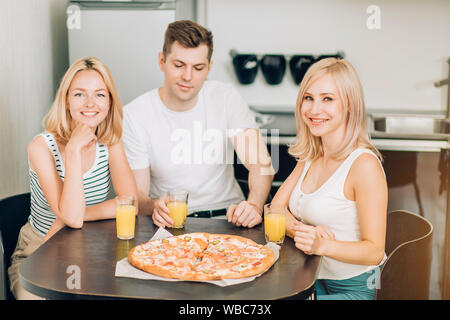 Due felice bella bionda caucasian giovani donne con il loro amico a mangiare una gustosa pizza e bere frullati di frutta, seduti a tavola in cucina, smili Foto Stock