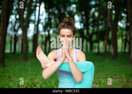 Thin brunette girl gioca sport ed esegue la bella e sofisticata yoga pone in un parco d'estate. Il verde lussureggiante foresta sullo sfondo. Donna facendo Foto Stock