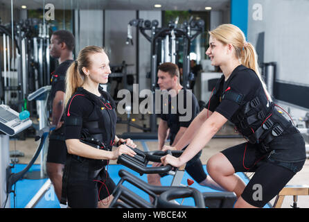 Due donne sorridenti vestiti giubbotti di EMS friendly parlando durante gli allenamenti in palestra per il fitness Foto Stock