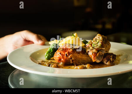 Primo piano di una persona che sta per servire un piatto di pollo arrosto con patate fritte e peperoni verdi come accompagnamento, in una cucina professionale Foto Stock