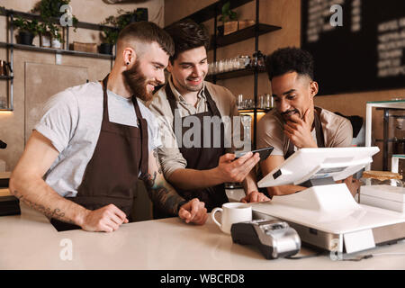 Tre uomini allegra baristi in piedi dietro il bancone presso la caffetteria, utilizzando il telefono cellulare Foto Stock