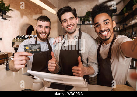 Tre uomini allegra baristi in piedi dietro il bancone presso la caffetteria, prendendo un selfie, mostra carta di credito Foto Stock