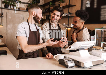 Tre uomini allegra baristi in piedi dietro il bancone presso la caffetteria, utilizzando il telefono cellulare Foto Stock