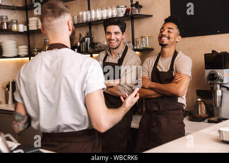 Il gruppo di allegro uomini baristi indossando grembiuli lavorando presso il contatore in cafe indoor, parlando Foto Stock