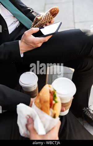 In prossimità di due giovani imprenditori che indossa abiti avente una pausa pranzo all'aperto presso le strade della città, a bere caffè e mangiando panini Foto Stock