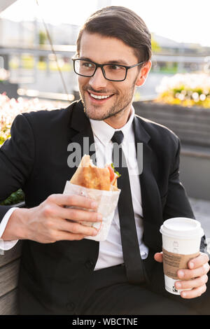 Attraente sorridente giovane impreditore indossa una tuta seduta su una panchina all'aperto presso la strada di città, avente la pausa pranzo Foto Stock