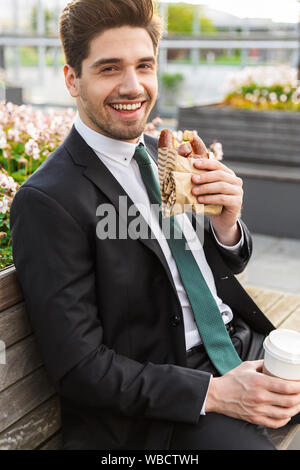 Attraente sorridente giovane impreditore indossa una tuta seduta su una panchina all'aperto presso la strada di città, avente la pausa pranzo Foto Stock