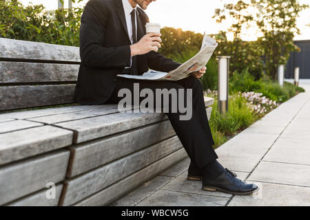Immagine ritagliata di un giovane imprenditore che indossa un abito seduta su una panchina all'aperto presso la strada di città, la lettura di un quotidiano mentre avente una pausa caffè Foto Stock
