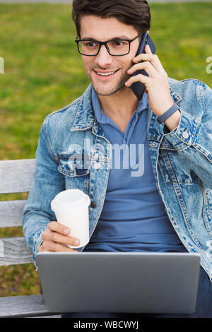 Bello sorridente giovane uomo vestito casualmente a trascorrere il tempo all'aperto presso la città, utilizzando il computer portatile mentre è seduto su una panchina e bere takeaway c Foto Stock