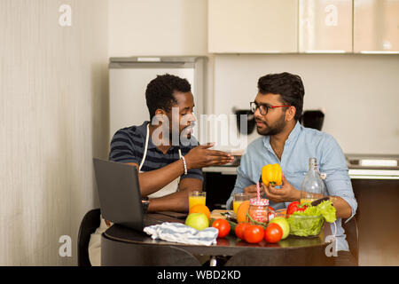 Due uomini in abiti casual seduta a tavola, amichevole parlare di qualcosa, preparare il frullato vegetale, il taglio di peperone, utilizzando il computer portatile ho Foto Stock