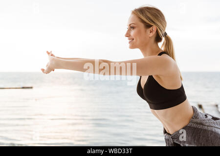 Immagine della ragazza bionda indossare tuta facendo esercizio mentre si lavora fuori sul molo vicino al mare al mattino Foto Stock