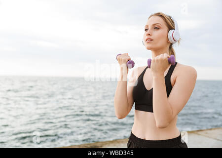 Immagine di serio carino donna che indossa tuta usando le cuffie mentre si lavora con i pesi sul molo vicino al mare al mattino Foto Stock