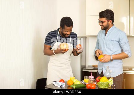 Indiano bello giovane uomo in bicchieri e americano africano uomo che indossa il grembiule divertendosi in cucina. Due amici maschi in piedi vicino alla scrivania con frutte e Foto Stock