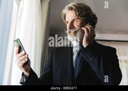Immagine di gioiosa imprenditore adulti indossando abito nero utilizza lo smartphone e auricolari mentre sta in piedi vicino alla finestra in hotel apartment Foto Stock
