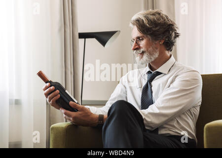Foto di un concentrato grigio senior con capelli business uomo seduto in poltrona al chiuso in casa tenendo la bottiglia con alcool bere vino. Foto Stock