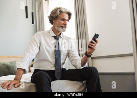 Foto di un grave concentrato grigio senior con capelli business uomo seduto sul letto all'interno di casa utilizzando il telefono cellulare. Foto Stock