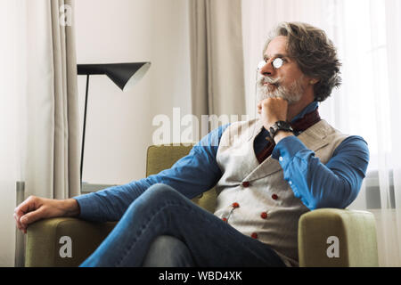 Immagine del fiducioso il professor adulto uomo elegante da indossare tuta e occhiali cercando in disparte mentre seduto sulla poltrona in hotel apartment Foto Stock