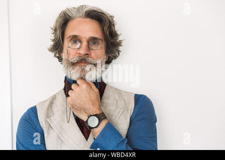 Immagine della barba lunga professore adulto uomo elegante da indossare tuta e occhiali guardando la fotocamera mentre in piedi su parete in appartamento Foto Stock