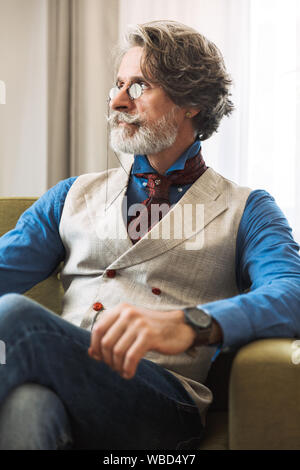 Immagine del Caucaso professore adulto uomo elegante da indossare tuta e occhiali cercando in disparte mentre seduto sulla poltrona in hotel apartment Foto Stock