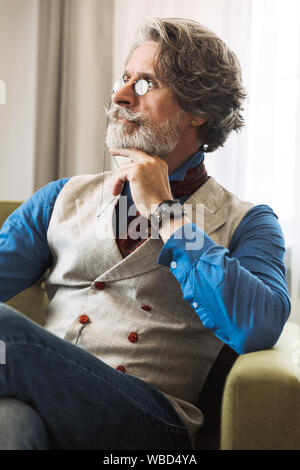 Immagine di grave professore adulto uomo elegante da indossare tuta e occhiali cercando in disparte mentre seduto sulla poltrona in hotel apartment Foto Stock