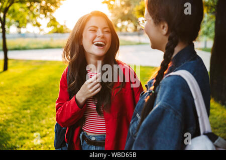 Immagine delle due ragazze ridere gli studenti indossando zaini parlando e guardando gli uni con gli altri mentre passeggiate nel parco verde Foto Stock