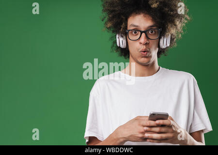 Foto Dettaglio di perplesso uomo con acconciatura afro guardando la fotocamera mentre si utilizza il cellulare e le cuffie isolate su sfondo verde Foto Stock