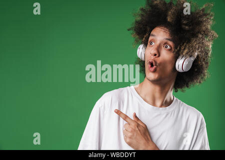 Immagine di un scioccato curly guy posa isolato sulla parete verde contesto l'ascolto di musica con cuffie di puntamento copyspace. Foto Stock