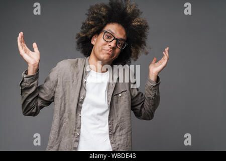 Foto di confuse giovane con acconciatura afro guardando la fotocamera e alzando le mani isolate su uno sfondo grigio Foto Stock