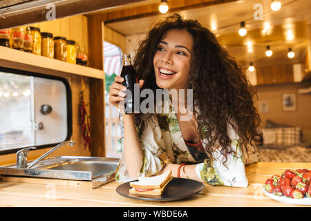 Ritratto di felice donna caucasica con ricci capelli lunghi a ridere e bere soda in accogliente cucina a rimorchio in ambienti interni Foto Stock