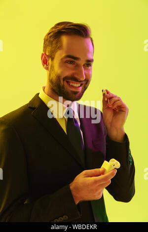 Bello sorridente giovane imprenditore indossare tuta formale in piedi isolato su sfondo giallo, scatola di contenimento con gli auricolari wireless Foto Stock