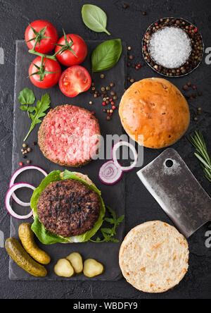 Carni alla brace e materie macinata di pepe hamburger di manzo su pietra tagliere con ciambelle di cipolla e pomodoro su sfondo nero. Il salato sottaceti e basilico. Parte superiore Foto Stock