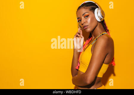 Immagine di athletic americano africano donna che indossa treccine afro ascolto di musica con cuffie isolate su sfondo giallo Foto Stock