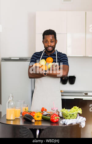 Attraente sorridente giovane barbuto dark scuoiati uomo che indossa il grembiule tenendo fresche e mature arance organico, mele e succo di pompelmo, rendendo frullato di frutta, godeteveli Foto Stock