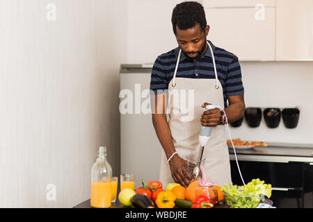 Giovani barbuto americano africano uomo che indossa polo t-shirt grembiule e la preparazione di un alimento sano, con blender per sminuzzare gli ingredienti, rendendo smo vegetale Foto Stock