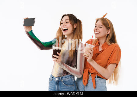 A piena lunghezza ritratto di due ragazze positive indossando abiti casual tenendo selfie foto e tenendo i bicchieri di plastica isolate su sfondo bianco Foto Stock