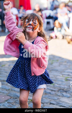 Bambino caucasico, ragazza, 5-7 anni, con dipinta la faccia di tiger, ballare saltando su e giù durante la outdoor evento musicale. Rivolta verso il visualizzatore, contatto visivo. Foto Stock