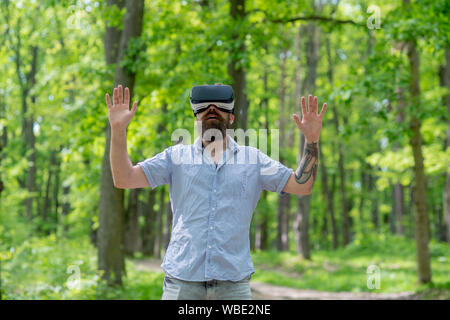 Uomo Barbuto indossando occhiali per realtà virtuale nella foresta. Lo smartphone utilizzando con occhiali VR auricolare. Uomo eccitato con la bocca aperta il roaming in boschi con Foto Stock