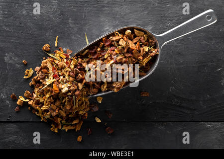 Cumulo di frutta secca di infusione del tè con arance e fragole miscelato con foglie di tè e un assortimento di erbe in metallo scoop su tavoli in legno nero backgroun Foto Stock