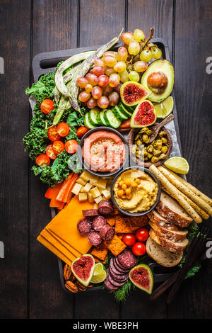 La carne e i formaggi antipasto. Salsicce, formaggio, hummus, verdure, frutta e pane su un vassoio nero, sfondo scuro. Foto Stock