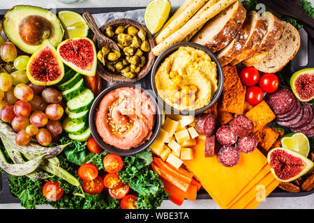 La carne e i formaggi antipasto. Salsicce, formaggio, hummus, verdure, frutta e pane su un vassoio nero, sfondo bianco. Foto Stock
