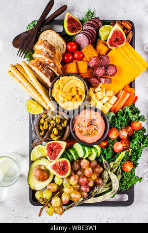 La carne e i formaggi antipasto. Salsicce, formaggio, hummus, verdure, frutta e pane su un vassoio nero, sfondo bianco. Foto Stock