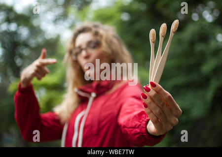 Ritratto di ragazza bionda con hipster occhiali in rosso scuro giacca tenendo un utile gli spazzolini da denti di bambù all'aperto nel parco urbano. Vivere Verde e Foto Stock