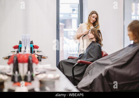 Bellezza, concetto di acconciatura, felice giovane donna e parrucchiere con capelli ferro rendendo pettinatura al salone di parrucchiere. Donna con capelli stilizzata parrucchiere Foto Stock