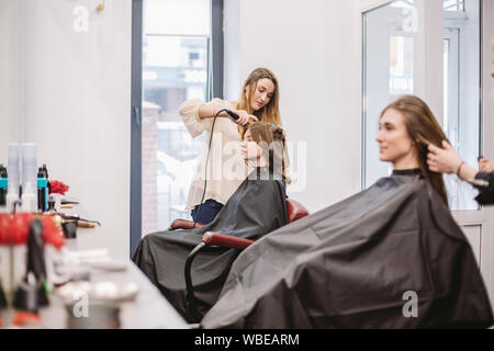 Bellezza, concetto di acconciatura, felice giovane donna e parrucchiere con capelli ferro rendendo pettinatura al salone di parrucchiere. Donna con capelli stilizzata parrucchiere Foto Stock