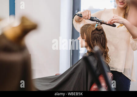 Bellezza, concetto di acconciatura, felice giovane donna e parrucchiere con capelli ferro rendendo pettinatura al salone di parrucchiere. Donna con capelli stilizzata parrucchiere Foto Stock