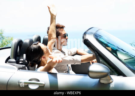 Giovani asiatici giovane in sella ad una autovettura convertibile su un lungomare, vista laterale Foto Stock