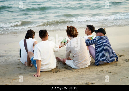 Un gruppo di giovani asiatici uomini adulti bere birra facendo clic su bottiglie tostare sulla spiaggia, vista posteriore Foto Stock