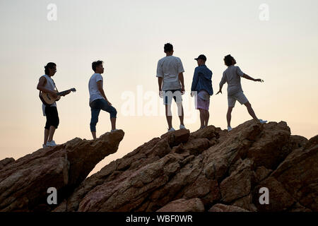 I membri di una rock band in piedi sulla parte superiore delle rocce in riva al mare a cantare a suonare la chitarra Foto Stock